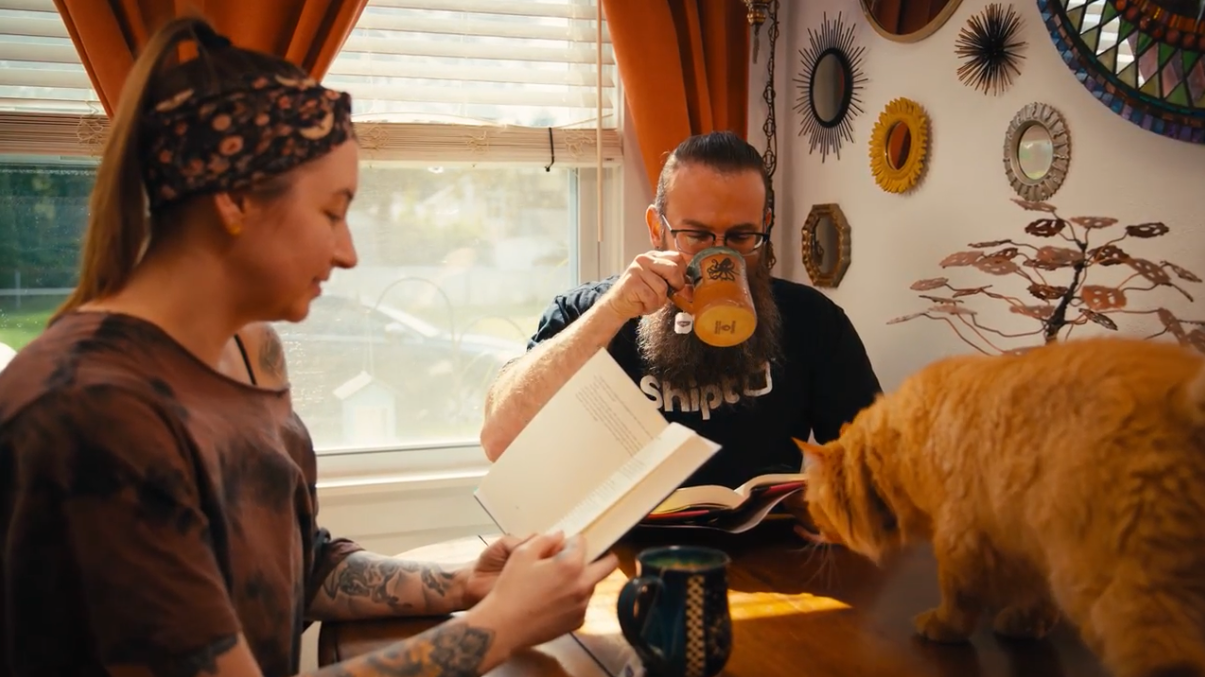 Two people sitting at a table, one is reading and the other drinking tea. An orange cat is standing on the table. 